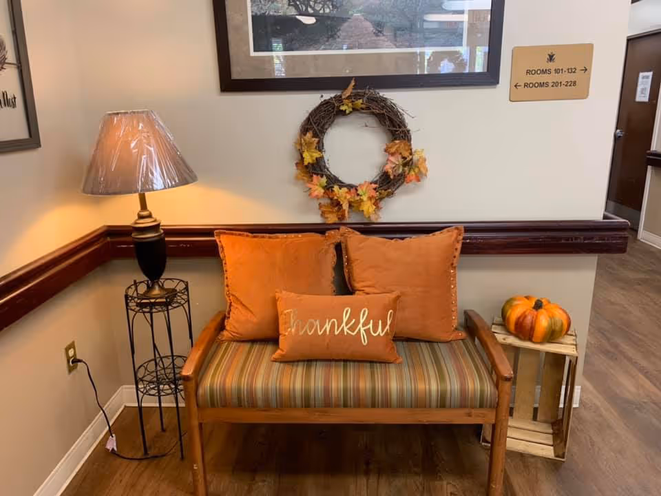 A cozy seating area with a wooden bench featuring striped upholstery and three orange cushions, one of which has the word 'thankful' embroidered on it. A decorative wreath with autumn leaves hangs on the wall above the bench. To the left, there is a floor lamp with a plastic cover on the shade, and to the right, a small wooden crate holds a decorative pumpkin. A sign on the wall indicates directions to rooms 101-132 and 201-228.