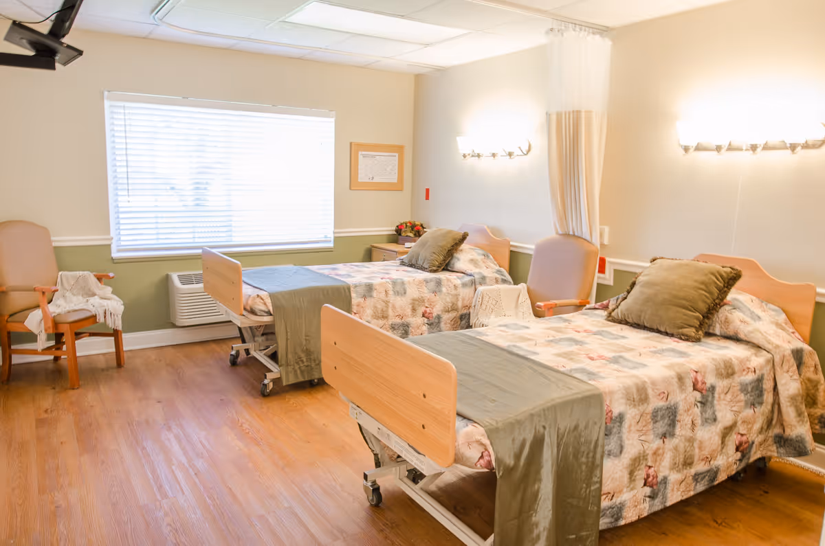 A bright room with two hospital-style beds, each with floral-patterned bedspreads and green throws. There are two beige chairs, one with a white knitted blanket draped over it. A large window with blinds lets in natural light, and the walls are painted light green and white. The floor is wooden, and there are wall-mounted lights above each bed.