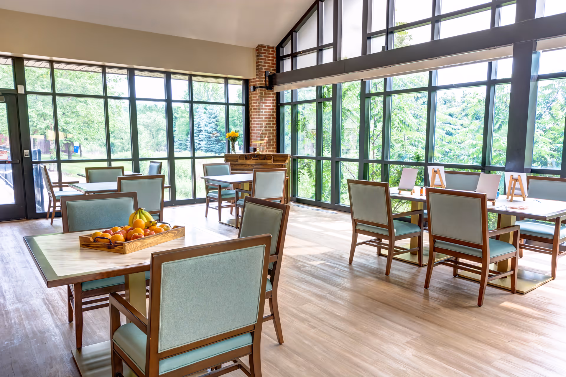 Bright dining room with large floor-to-ceiling windows letting in natural light, several wooden tables with light blue cushioned chairs, a tray of fruit on one table, and a vase with yellow flowers on a wooden side table near a brick column.