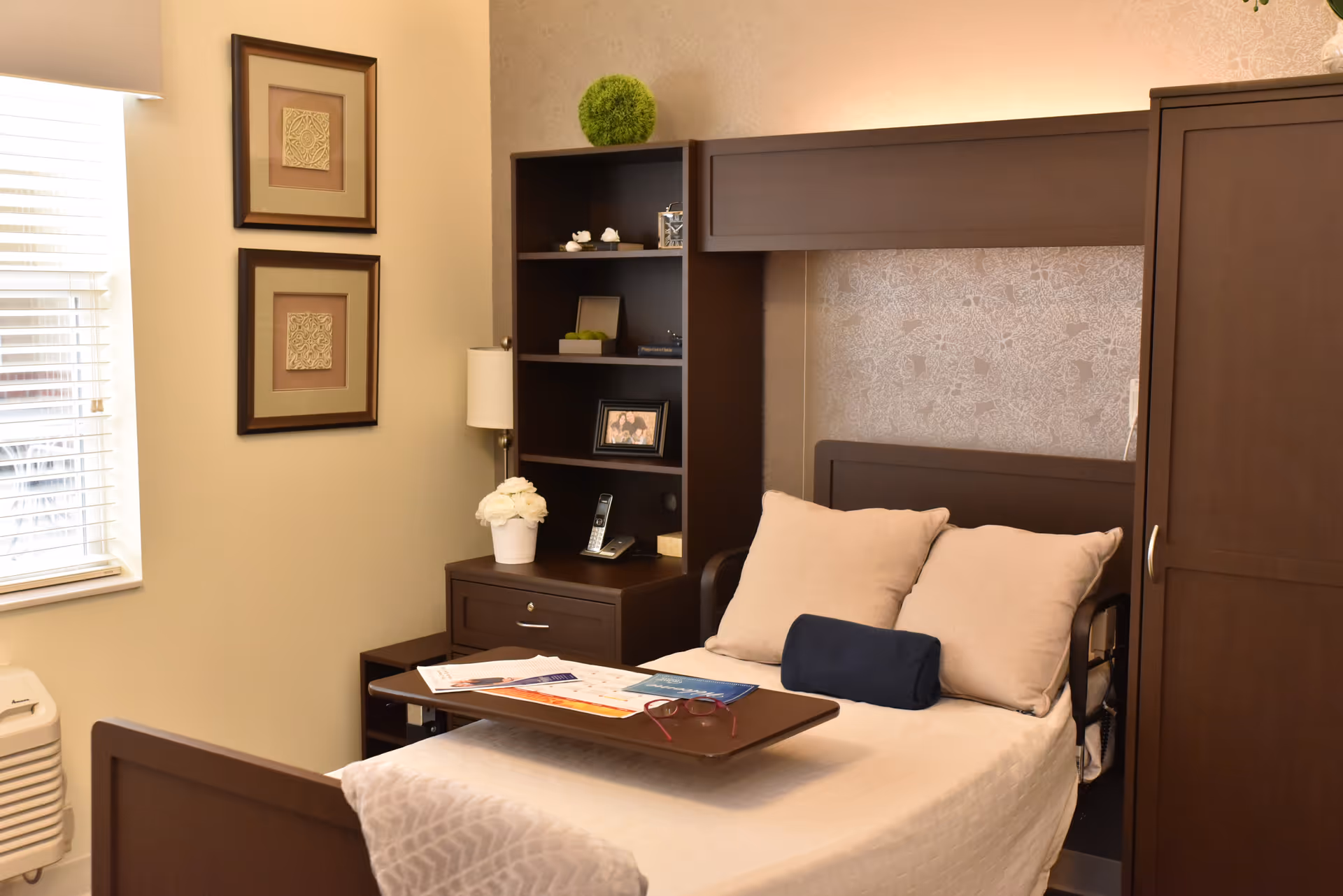 A neatly arranged bedroom in a senior living facility featuring a single bed with beige pillows and a blue bolster pillow. The bed has a wooden headboard with built-in shelving and a side table holding a lamp, a white flower pot, and a cordless phone. There are two framed artworks on the wall and a window with blinds letting in natural light.