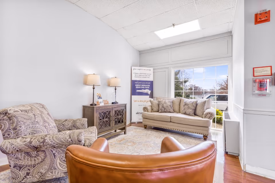 A bright and cozy living room area with a beige sofa adorned with patterned cushions, a paisley-patterned armchair, and a brown leather chair. There is a wooden sideboard with two lamps on it, a large window letting in natural light, and a standing sign with guest services information. The room has light-colored walls and a patterned rug on the wooden floor.