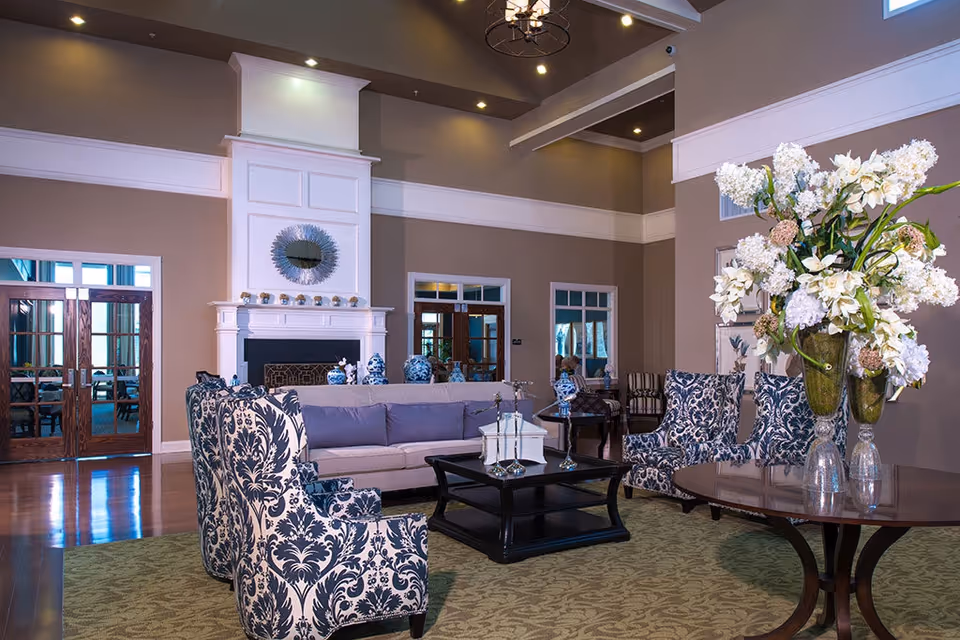 Spacious communal living room with patterned armchairs and a sofa around a coffee table, a fireplace in the background, and a large floral arrangement on a side table.