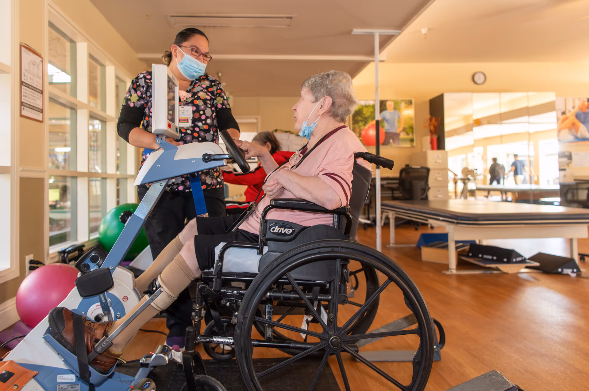 A healthcare worker wearing a mask assists an elderly woman in a wheelchair who is using a leg exercise machine in a rehabilitation center room with exercise equipment and large windows.