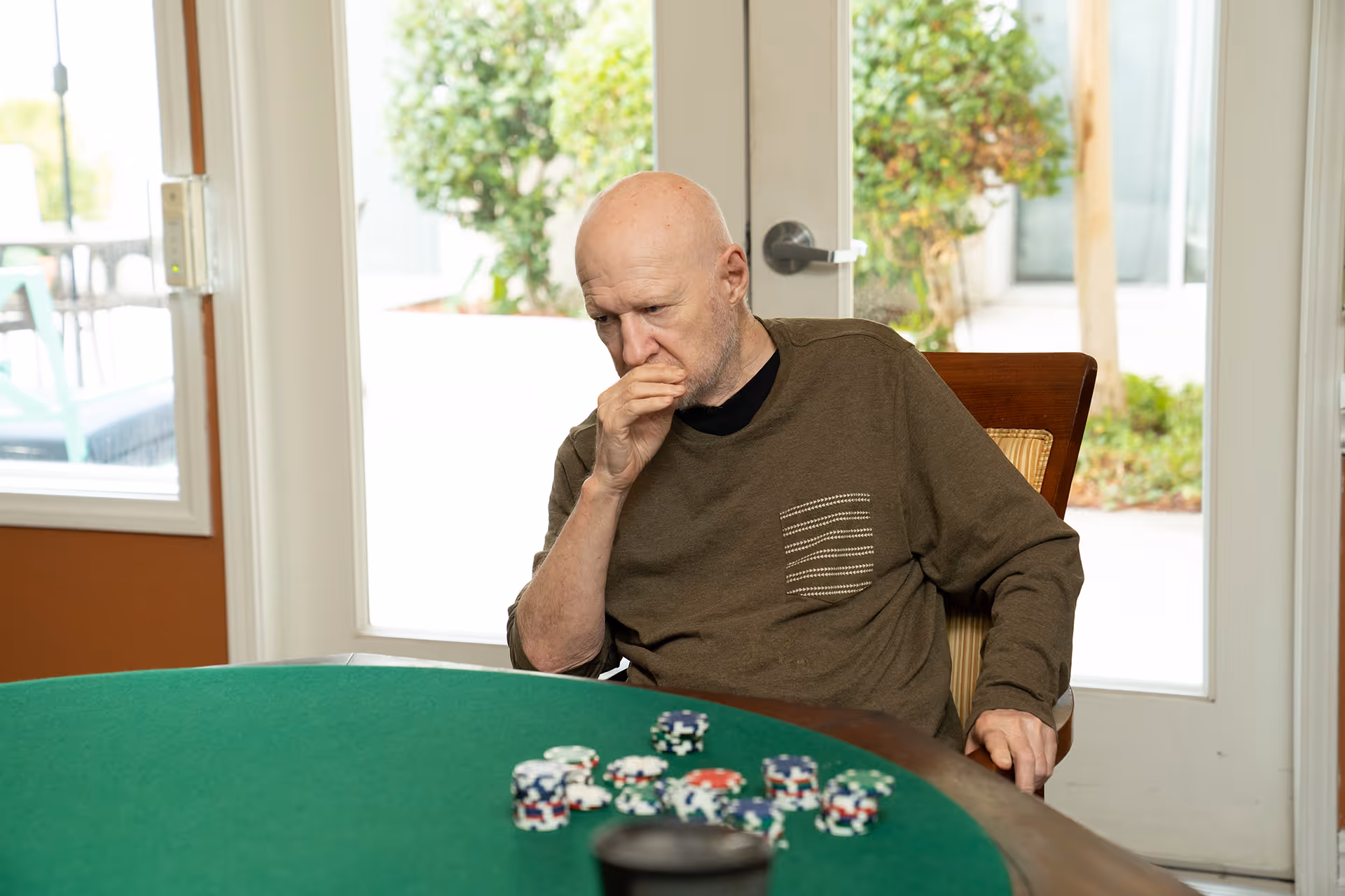 An elderly man sitting at a green poker table indoors, looking thoughtful with his hand near his mouth. There are poker chips scattered on the table. Behind him is a glass door with a view of greenery outside.