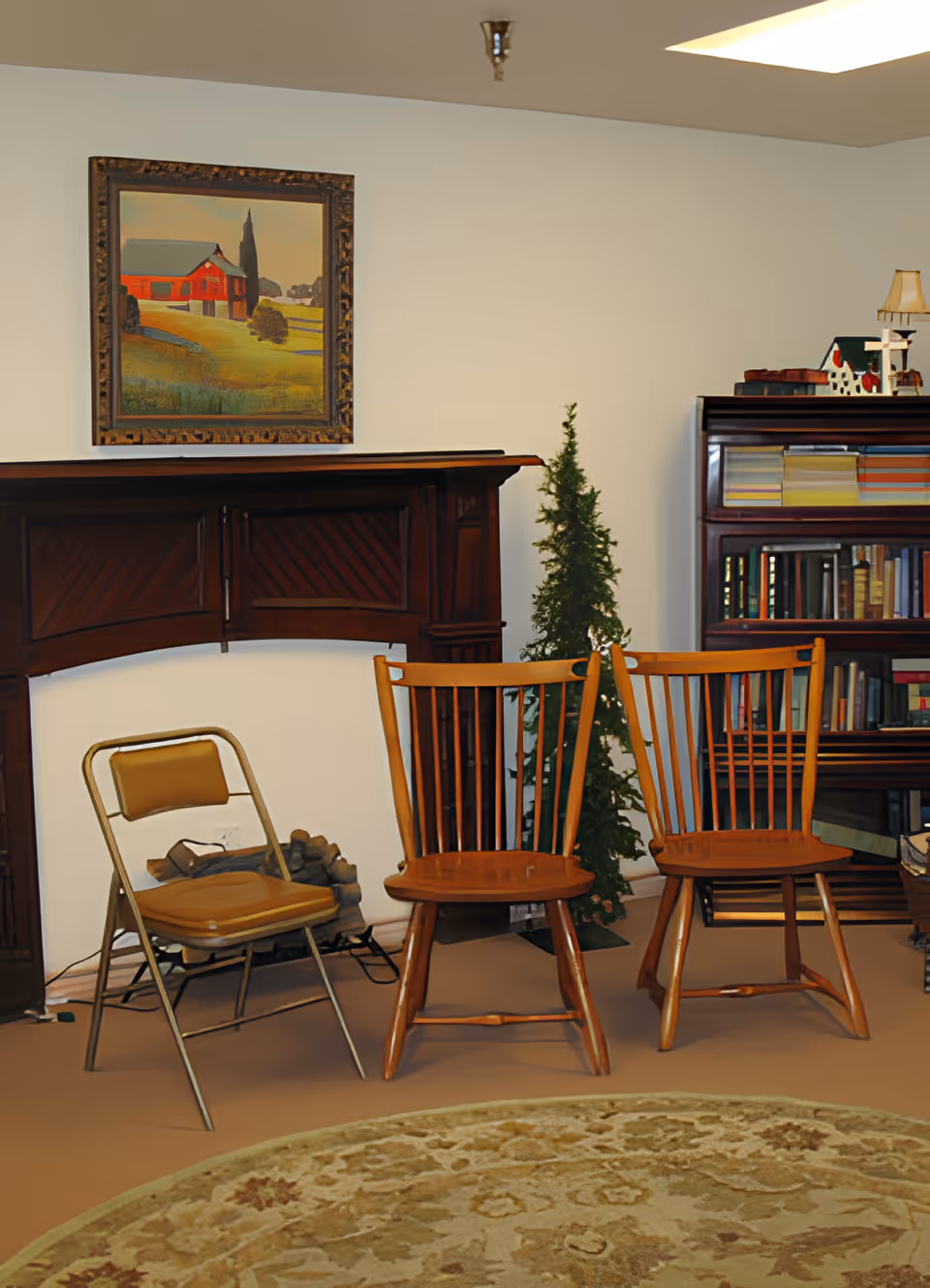 A cozy interior room with three chairs arranged in front of a wooden fireplace mantel. Two wooden chairs and one metal folding chair with a padded seat are visible. A small artificial Christmas tree is placed between the chairs and a wooden bookshelf filled with books is in the background. A framed painting of a red barn hangs above the fireplace mantel.