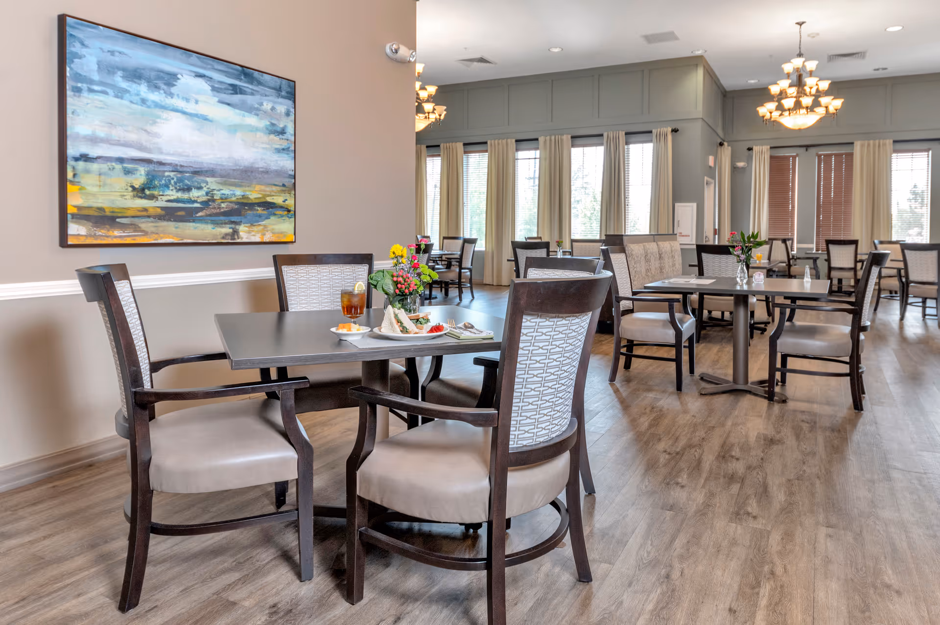 A bright dining room with several tables and chairs arranged neatly. Each table has a small flower vase, and one table in the foreground has a plate of food and a glass of iced tea. Large windows with cream-colored curtains allow natural light to fill the room. A large abstract painting hangs on the beige wall to the left, and elegant chandeliers hang from the ceiling.