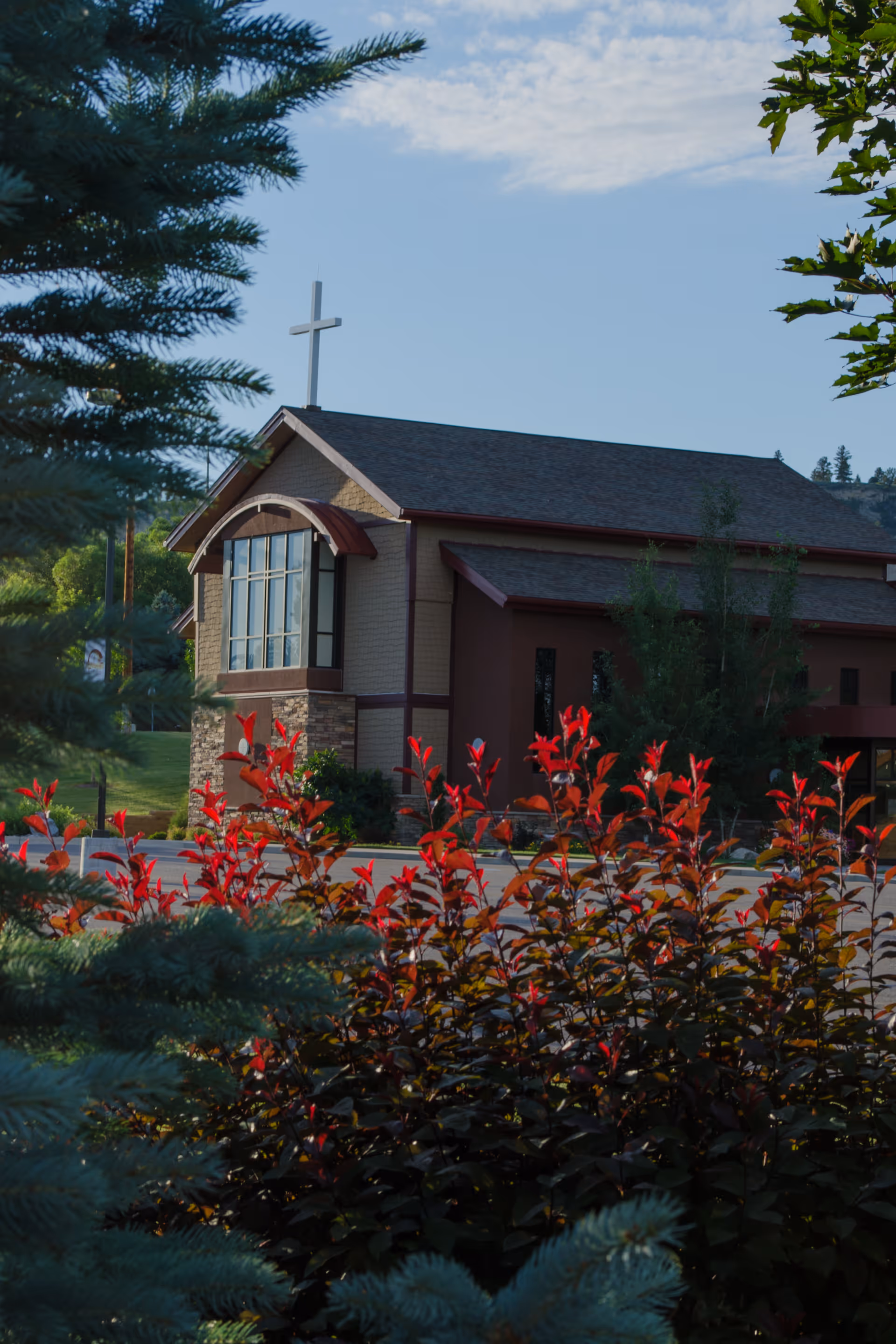 Exterior view of a building with a cross on the roof, partially obscured by green and red foliage in the foreground under a blue sky with some clouds.
