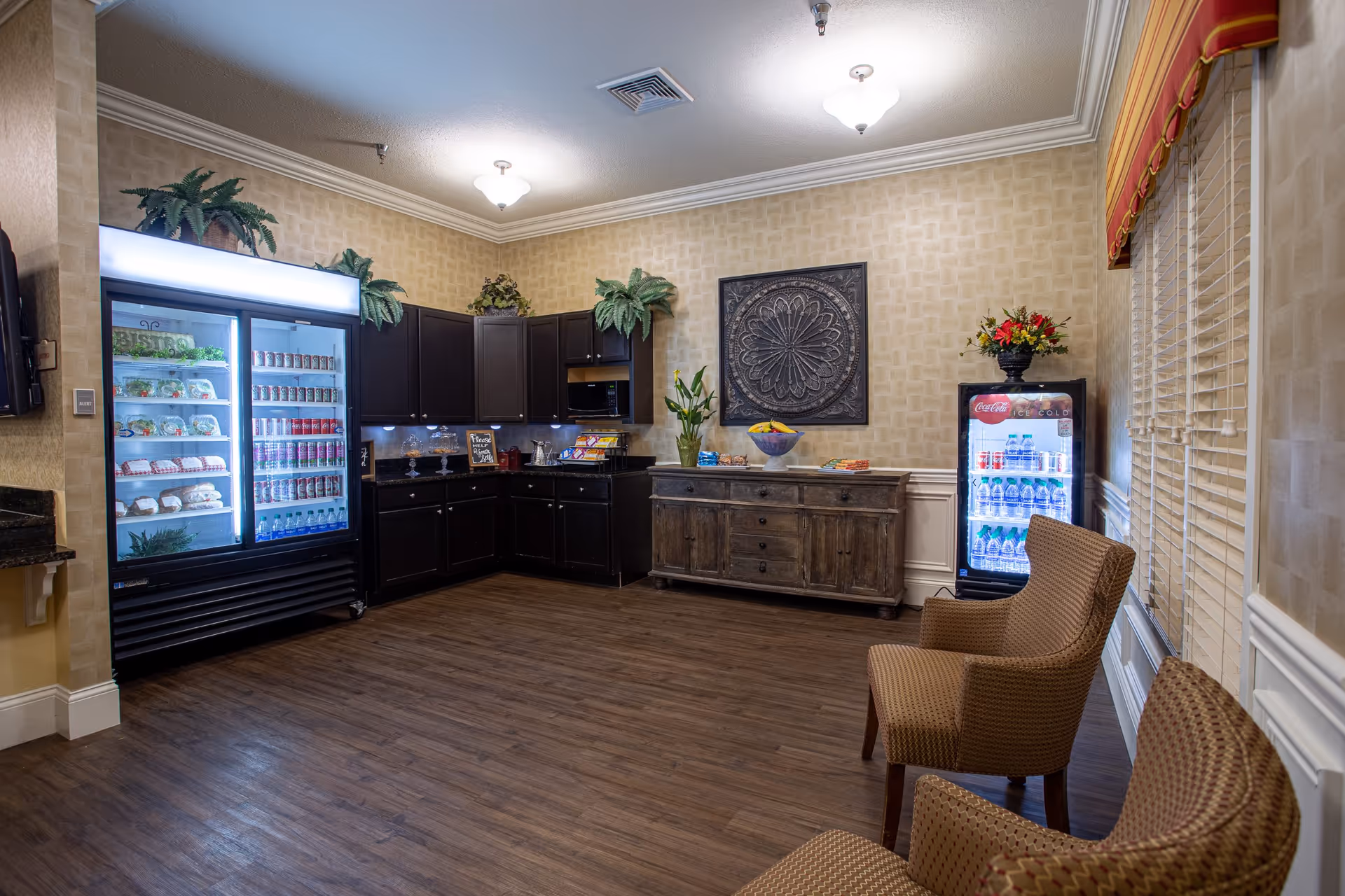 A senior living facility snack area with refrigerated display cases, dark cabinets, a wooden sideboard, and seating chairs.