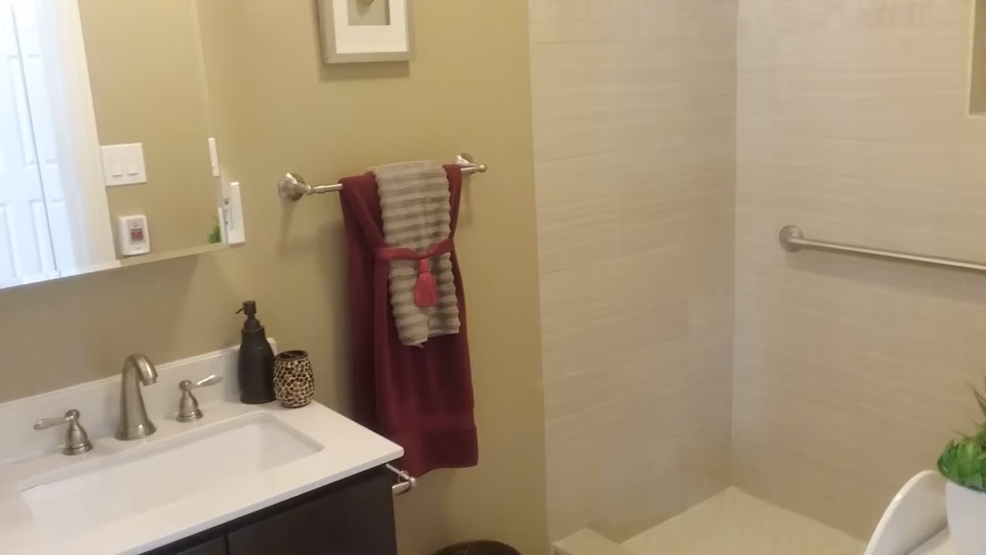 A bathroom with a white sink and silver faucet, a soap dispenser and a decorative cup on the counter. A towel rack on the wall holds a folded red towel with a smaller gray towel on top. The shower area has beige tiled walls and a silver grab bar. A small green plant is visible on the right side.
