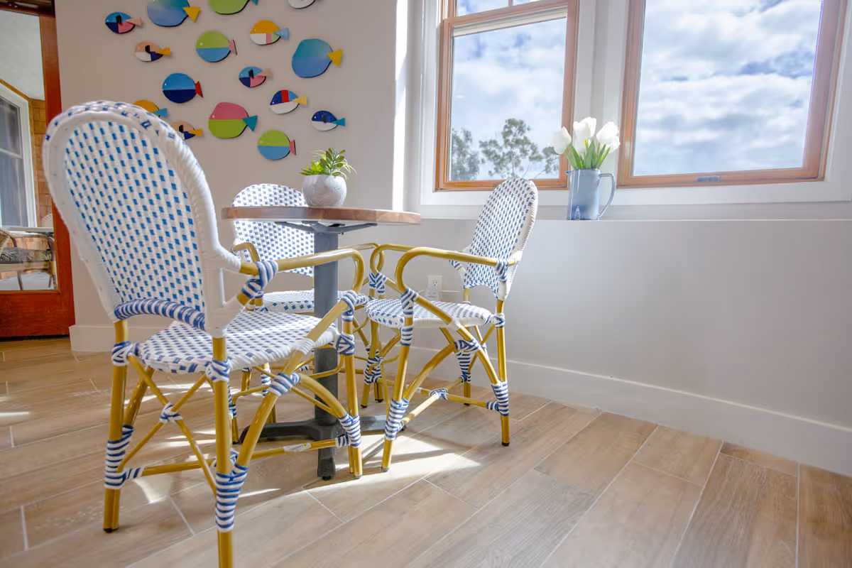 A bright room with a small round table and three wicker chairs with blue and white woven seats and backs. A potted plant sits on the table. On the wall behind the table, there is colorful fish-shaped wall art. A large window lets in natural light, with a vase of white flowers on the windowsill. The floor is light wood tile.