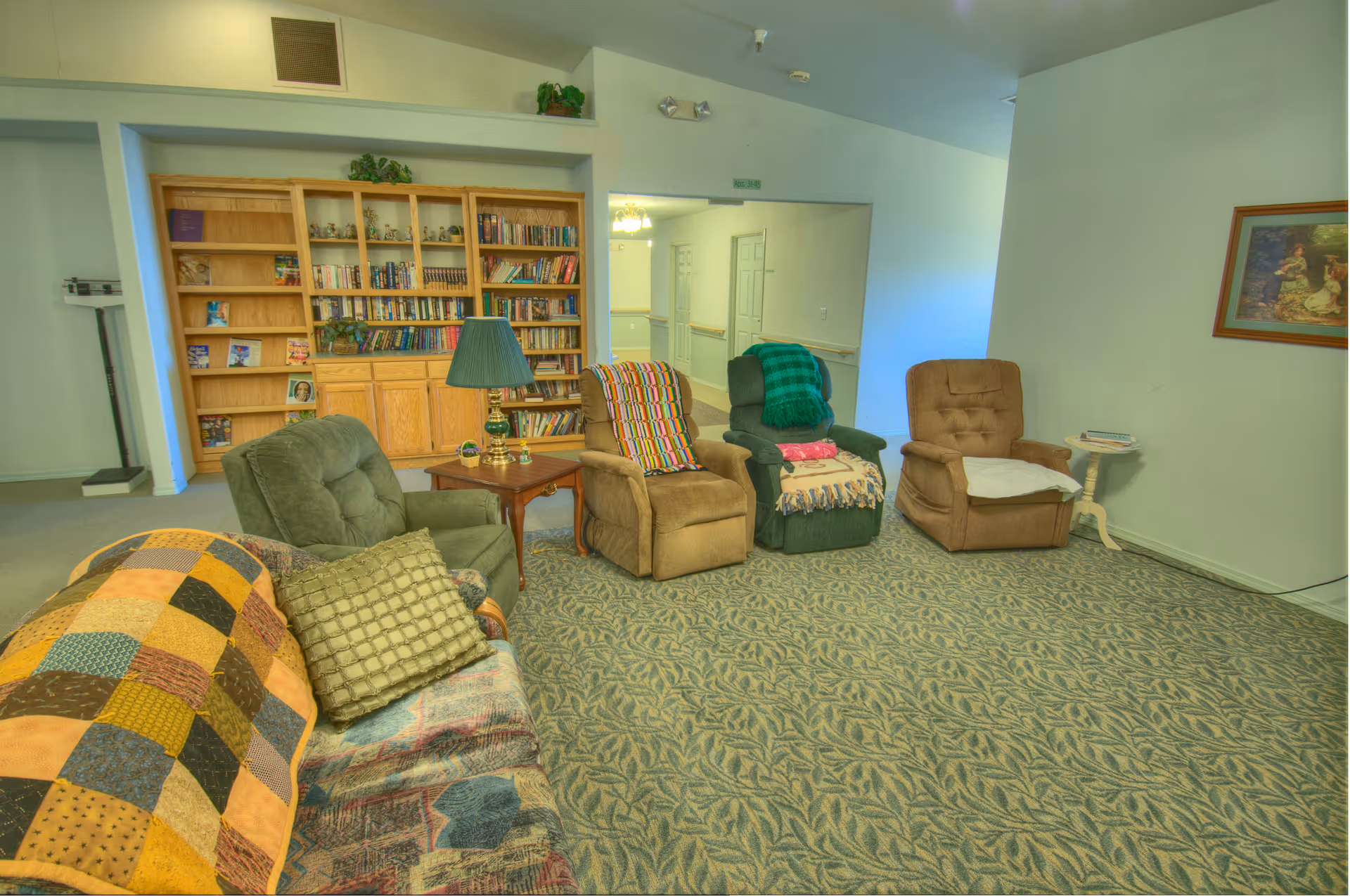 A cozy living room area in an assisted living facility featuring a patterned carpet, a couch with a patchwork quilt and pillows, three recliner chairs each with a throw blanket, a wooden side table with a lamp, and a large wooden bookshelf filled with books and decorative items. A framed picture hangs on the wall and a hallway is visible in the background.