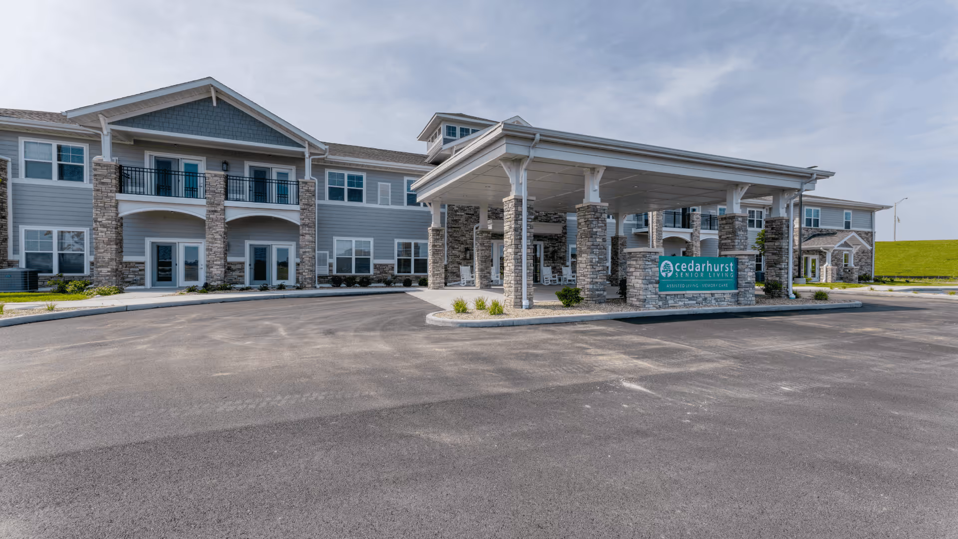Exterior front view of Cedarhurst Senior Living of McHenry building with a covered entrance supported by stone pillars and a sign displaying the facility name. The building is two stories with multiple windows and a paved driveway in front.