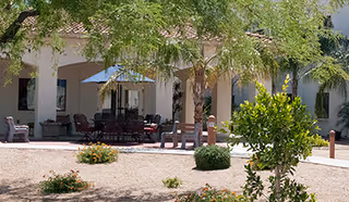 Outdoor patio area with tables, chairs, and umbrellas surrounded by desert landscaping including small bushes and trees in front of a building with arches and a tiled roof.