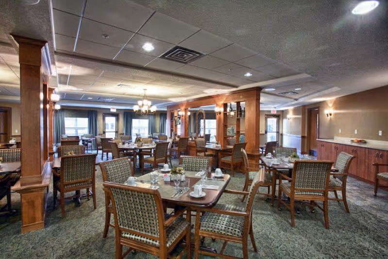 A spacious dining room with multiple wooden tables and cushioned chairs arranged neatly. Each table is set with white cups, saucers, glasses, and small flower centerpieces. The room features carpeted flooring, wood-paneled columns, and warm lighting from ceiling fixtures and wall sconces. Large windows with curtains allow natural light into the space.