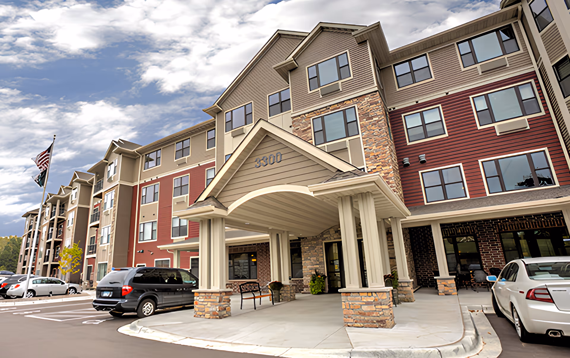 Exterior view of a multi-story senior living facility building with a covered entrance supported by columns. Several parked cars are visible in front of the building, and an American flag is flying on a flagpole near the entrance. The sky is partly cloudy.