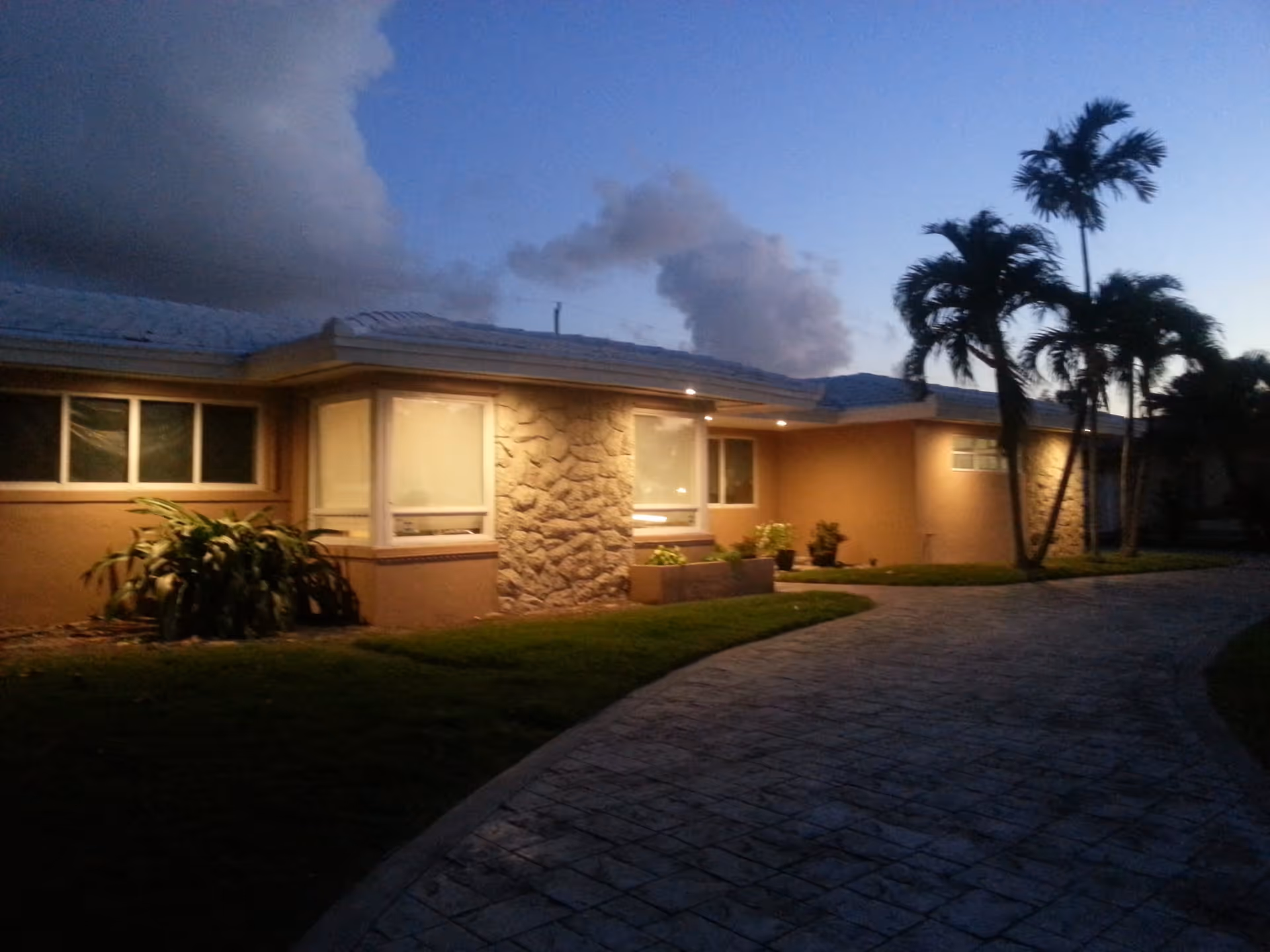 Exterior view of a single-story building at dusk with illuminated windows, stone and stucco walls, a curved paved driveway, and several palm trees in the yard under a partly cloudy sky.