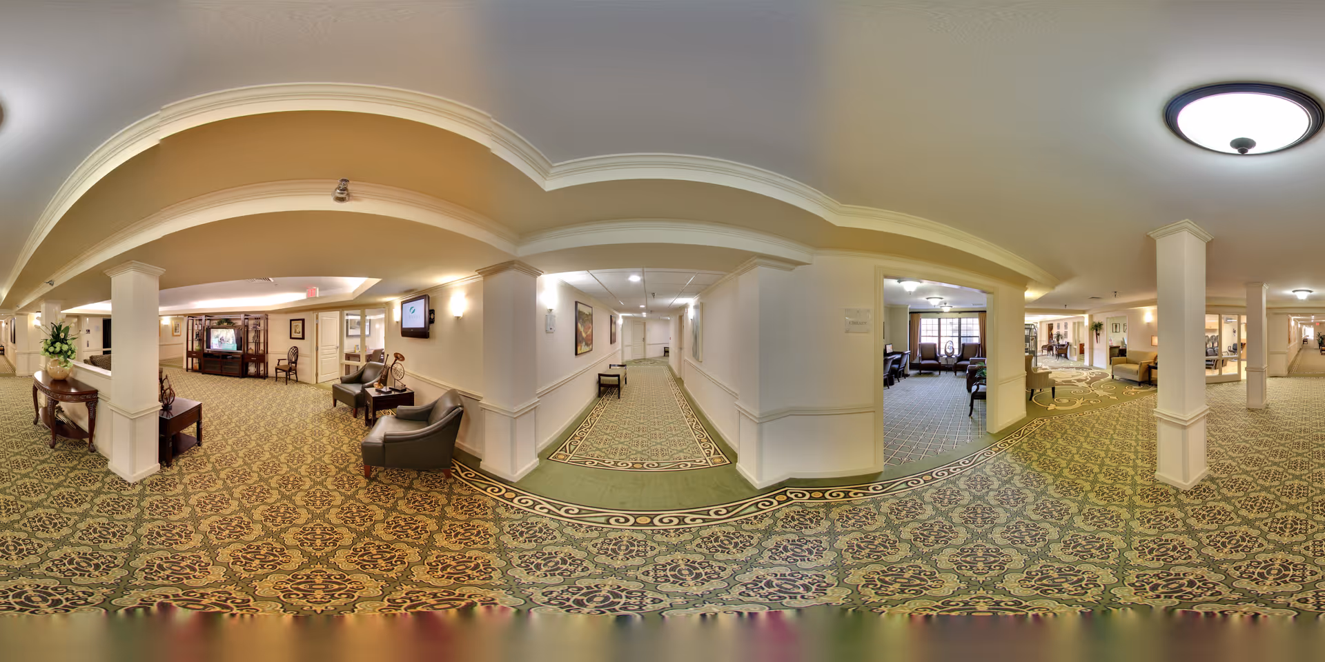 A spacious senior living community interior hallway with patterned carpet, white walls, and ceiling lights. The area includes seating arrangements with chairs and tables, decorative plants, and framed artwork on the walls. Multiple corridors and rooms are visible, creating an inviting and comfortable atmosphere.