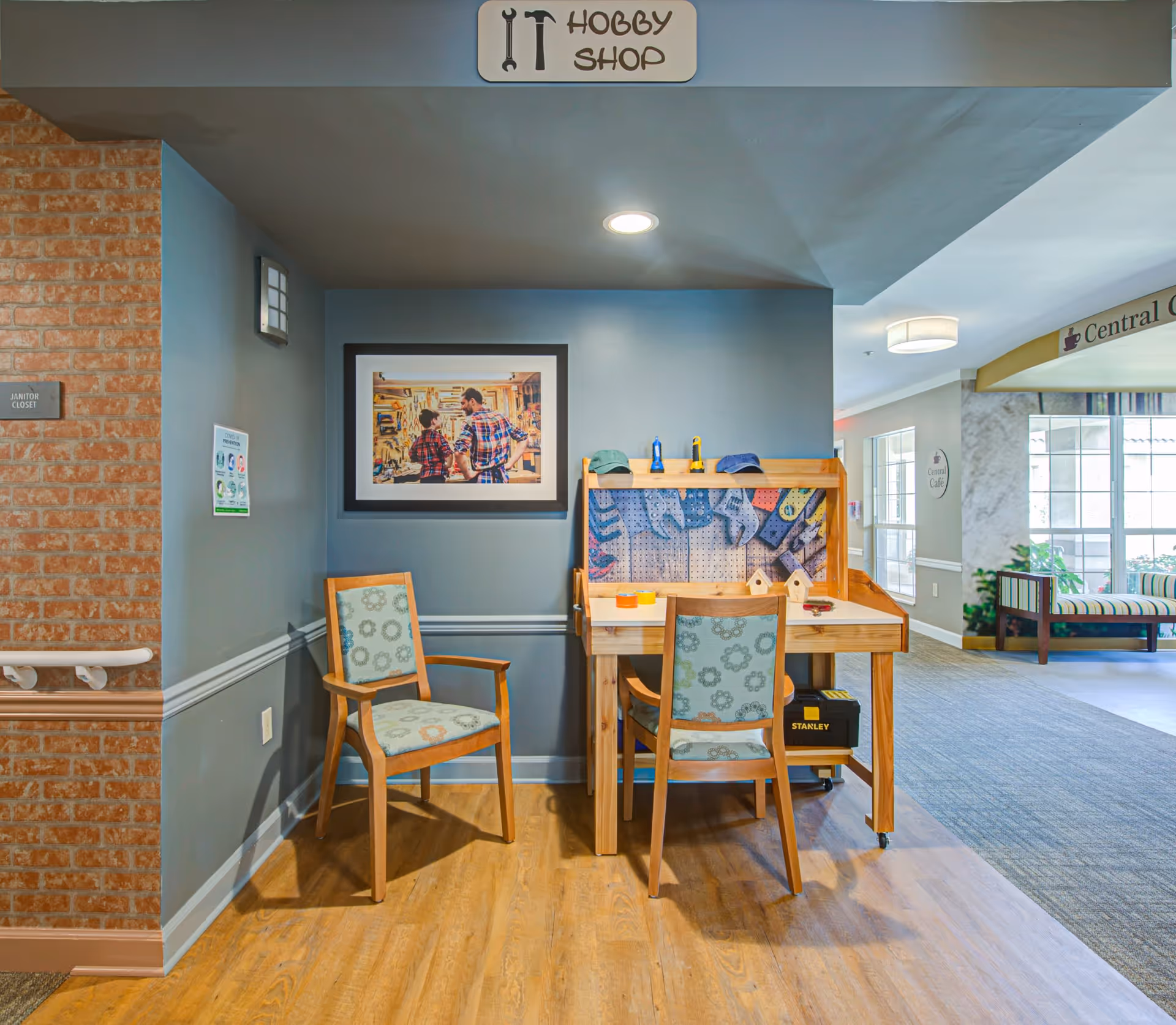 A hobby shop area in a senior living community featuring a wooden workbench with tools and two chairs with floral upholstery. Above the workbench is a sign that reads 'Hobby Shop'. On the wall behind the workbench is a framed picture of two people in a workshop. The surrounding area includes a brick wall with a janitor closet sign and a hallway leading to a seating area with large windows.