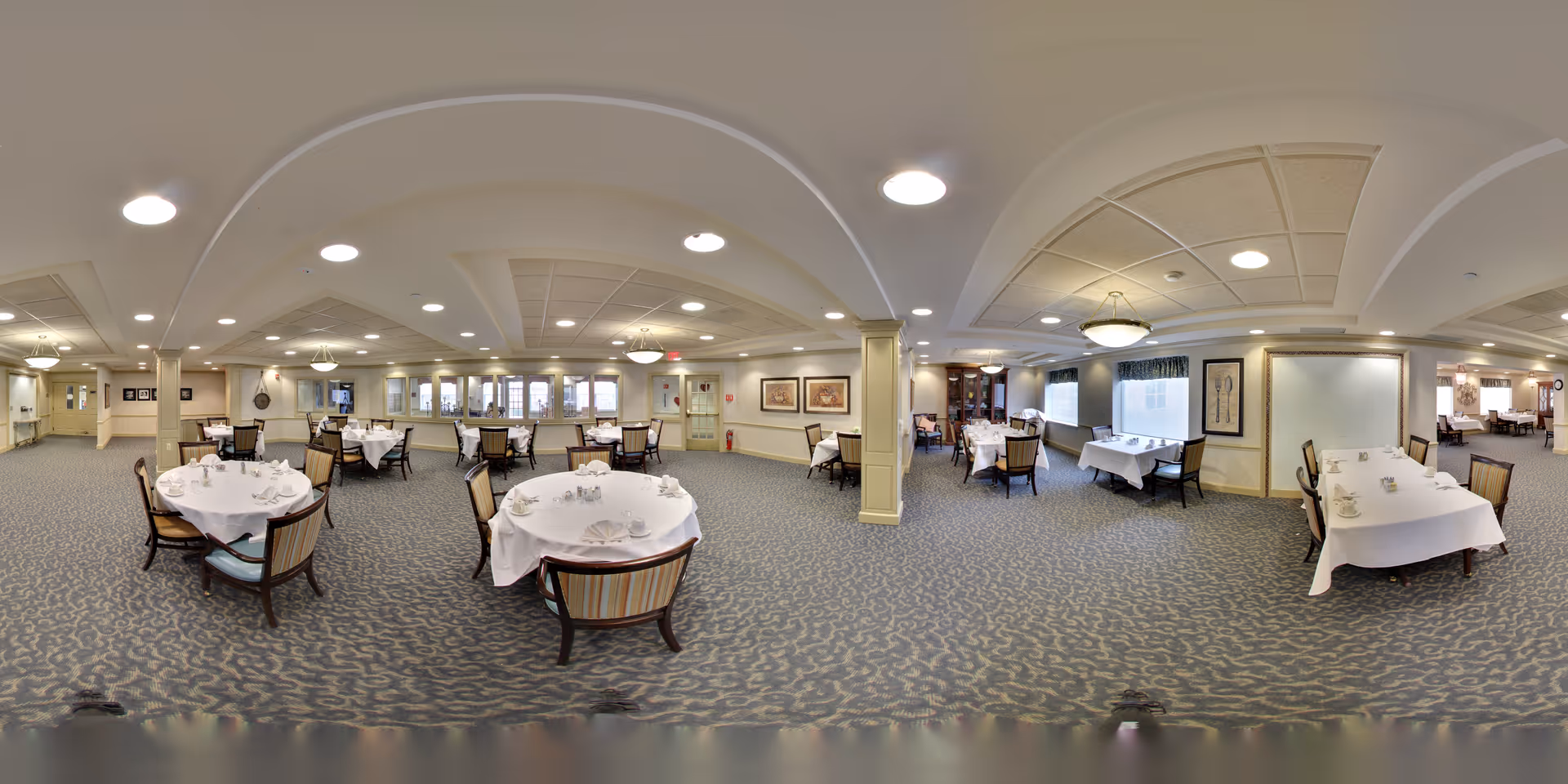 Large dining room with multiple round tables set with white tablecloths and chairs in a senior living facility.