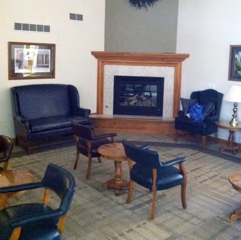 A cozy living room area with a fireplace centered on a wall. The room features several dark blue upholstered chairs and a loveseat arranged around small wooden tables. There are framed pictures on the walls and a table lamp on a side table to the right of the fireplace.