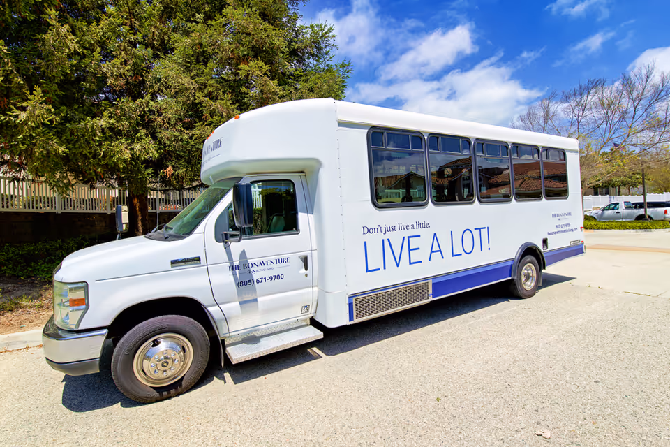 White senior-living shuttle bus parked outdoors with "Don't just live a little. LIVE A LOT!" printed on its side.