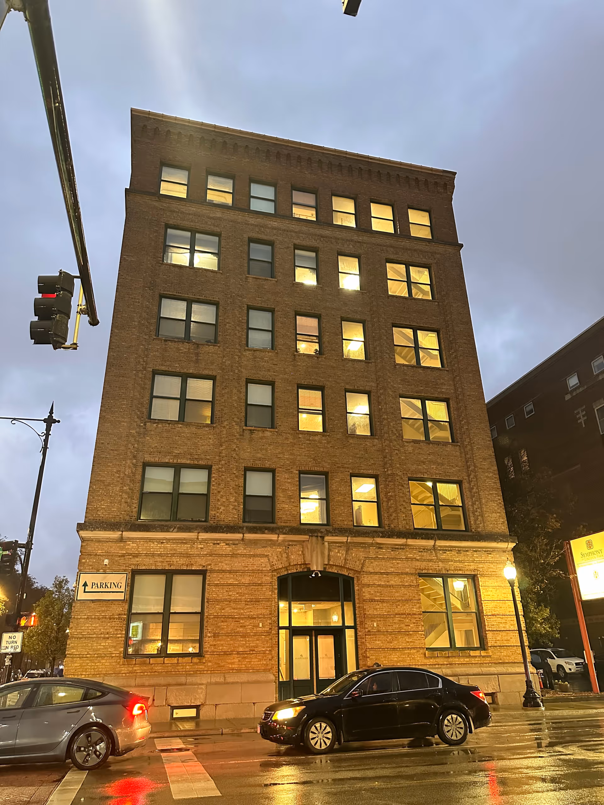 A multi-story brick building at dusk with illuminated windows. Two cars are stopped at a traffic light in front of the building on a wet street. A parking sign is visible on the building's left side, and a street lamp is on the right side near a signboard.
