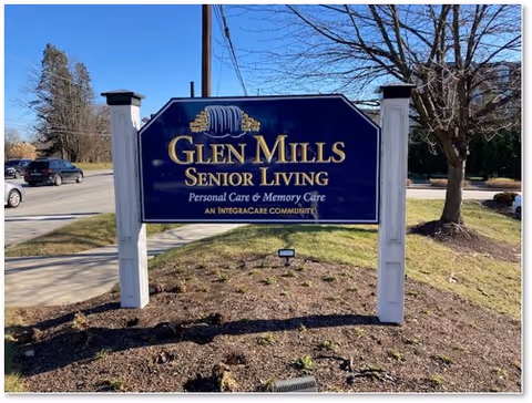 A blue entrance sign on white posts reading "Glen Mills Senior Living" on a grassy roadside lawn.