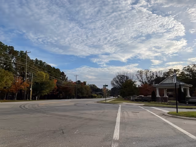 A wide street with a curve, bordered by trees with autumn foliage on the left and a small pavilion structure on the right under a partly cloudy sky.