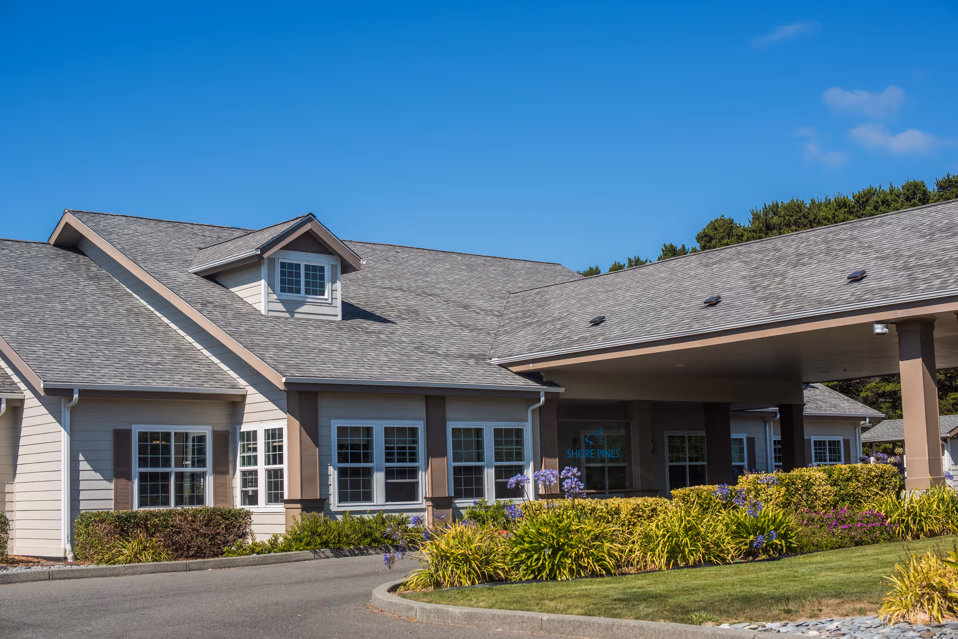 Front exterior of Shore Pines Senior Living building with a covered entrance, windows, and landscaped flower beds under a clear blue sky.