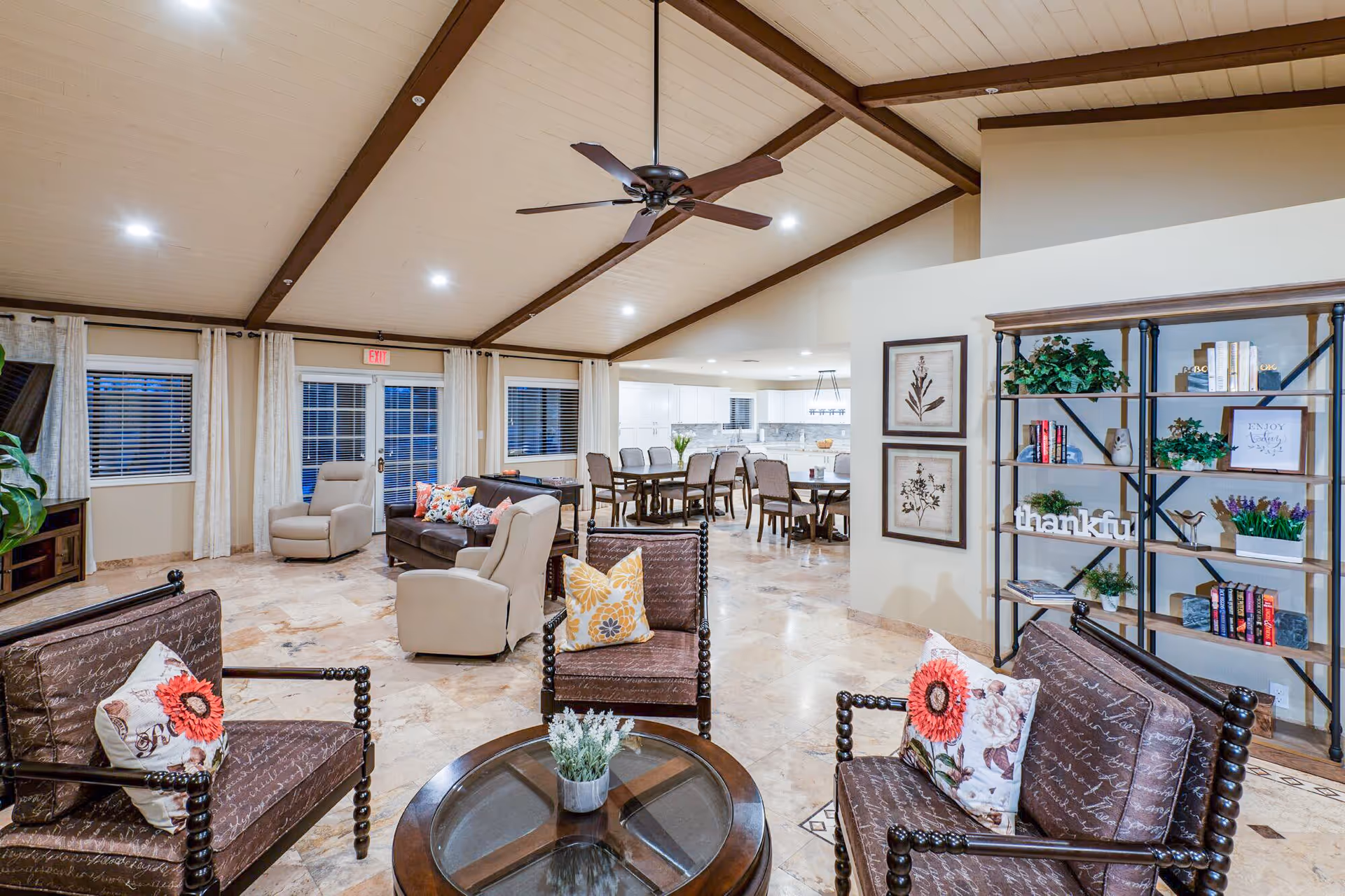 Spacious and well-lit senior living common area with a vaulted ceiling and exposed wooden beams. The room features multiple seating options including cushioned armchairs with floral pillows, a dark brown sofa, and beige recliners. A round glass-top coffee table with a small plant is in the center. In the background, there is a dining area with several tables and chairs, and a modern kitchen with white cabinetry and a marble backsplash. Decorative shelving with plants, books, and framed artwork adorns one wall.