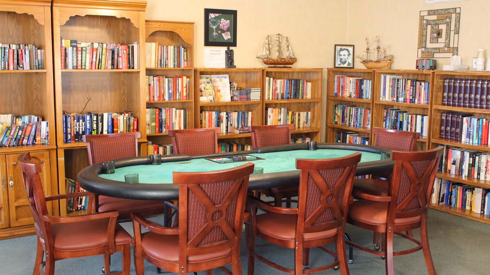 A room with wooden bookshelves filled with books and decorative items, including model ships and framed pictures. In the center, there is a green felt poker table surrounded by eight wooden chairs with red cushioned seats and backs.
