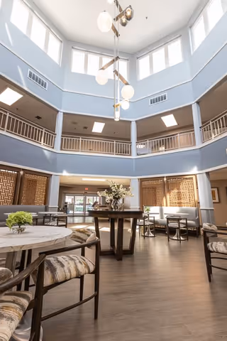 Bright and spacious two-story common area with large windows near the ceiling allowing natural light to fill the space. The room features a modern chandelier hanging from the ceiling, round tables with cushioned chairs, and a central table decorated with a floral arrangement. The walls are painted light blue with white trim, and there is a second-floor balcony railing overlooking the area.