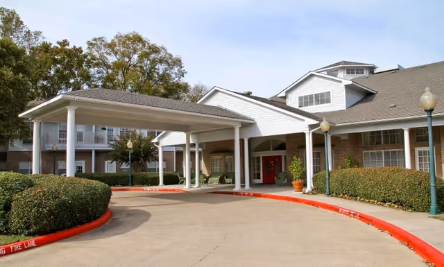 Front exterior view of a senior living facility with a covered entrance supported by white columns, red double doors, trimmed bushes, and street lamps along the driveway.
