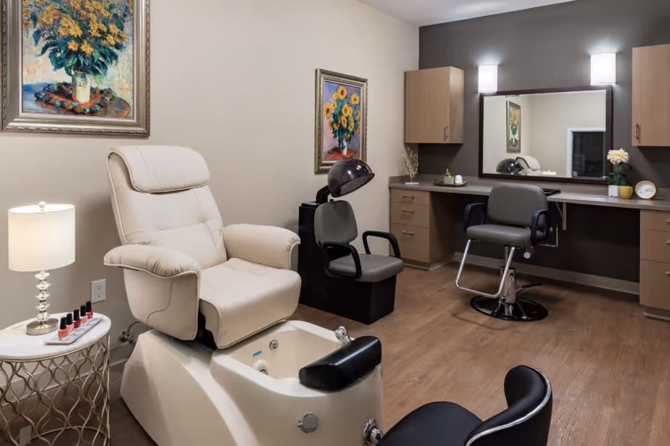 A well-lit salon room with a beige pedicure chair and foot bath, a gray salon chair in front of a large mirror, a hair dryer chair, wooden cabinets, and two framed paintings of sunflowers on the walls. A small side table with a lamp and nail polish bottles is next to the pedicure chair.