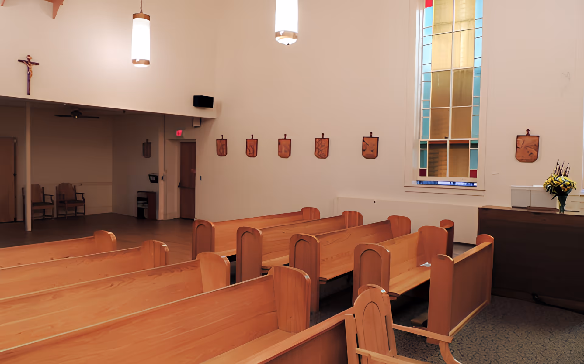 Interior view of a chapel with wooden pews arranged in rows, a stained glass window, religious plaques on the wall, a crucifix, and a small table with a flower arrangement.