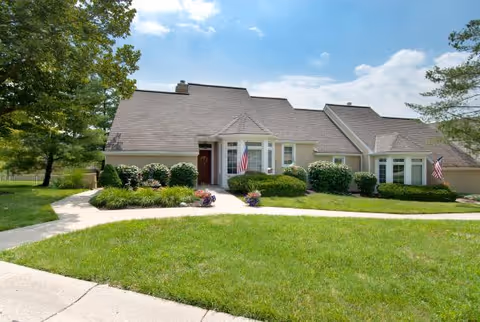 Exterior view of a single-story residential building with beige siding and a shingled roof, surrounded by green grass, bushes, and trees under a partly cloudy sky. Two American flags are displayed near the entrances.