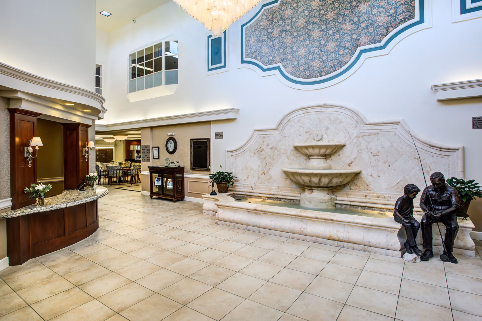 Spacious senior living facility lobby with a large decorative indoor fountain featuring statues of an elderly man and a child fishing. The area has tiled floors, a wooden reception desk with granite countertop, wall sconces with lamps, and a seating area visible in the background. The walls are adorned with floral wallpaper and a large chandelier hangs from the ceiling.
