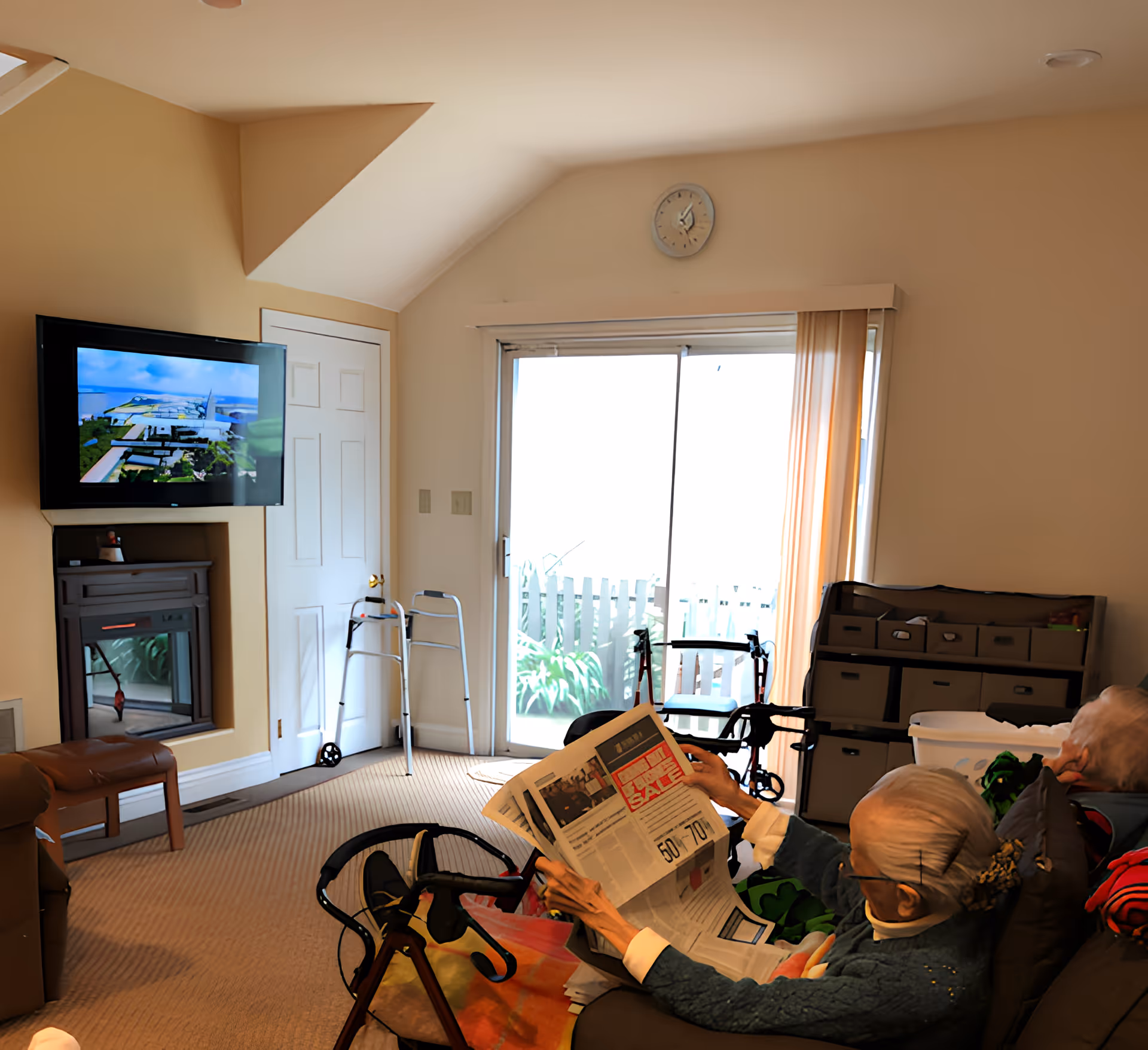 A cozy living room in a senior living facility with two elderly individuals seated on a couch. One person is reading a newspaper while the other looks on. The room features a wall-mounted TV, a fireplace, a walker, and a sliding glass door with vertical blinds leading outside. There is a clock on the wall above the door and storage bins against the wall.
