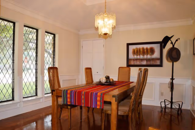 A dining room with a wooden table covered by a colorful striped table runner. Six wooden chairs surround the table. Three tall windows with diamond-patterned glass are on the left wall, letting in natural light. A chandelier hangs above the table. On the right wall, there is a framed painting of trees and a coat rack holding two hats.