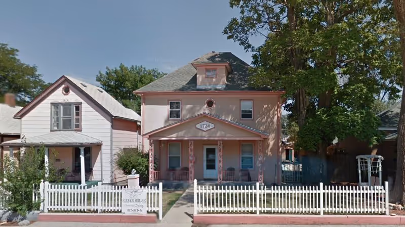 Front exterior view of a two-story pink house with a small porch and white picket fence. There is a sign in front of the fence that reads 'Eddie's House'. The house is flanked by another smaller house on the left and large trees on the right.