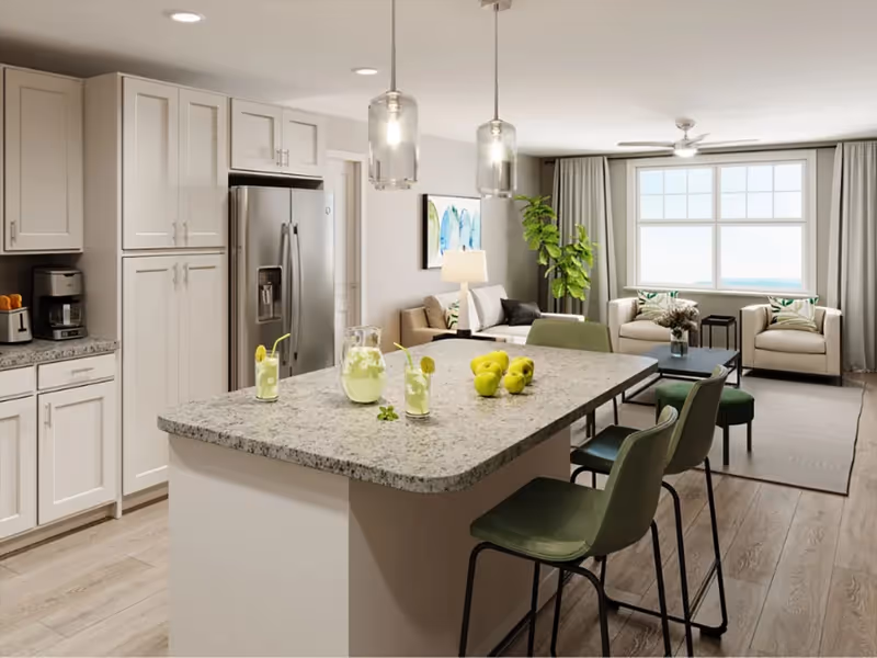 A modern open-concept kitchen and living room area. The kitchen features white cabinetry, a stainless steel refrigerator, a coffee maker, and a large granite island with three green bar stools. On the island, there is a pitcher and glasses of lemonade and a small pile of green apples. The living room has a large window with curtains, two beige armchairs, a beige sofa, a coffee table with a flower arrangement, a floor lamp, and a large green plant. The space is bright and well-lit with pendant lights above the island and recessed ceiling lights.