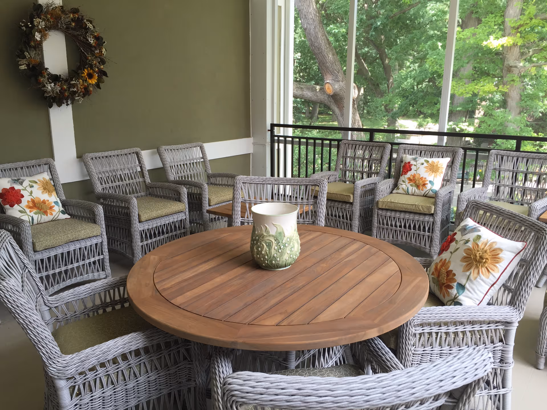 A cozy outdoor seating area with a round wooden table surrounded by gray wicker chairs with green cushions and floral pillows. A decorative wreath hangs on the olive green wall, and large windows reveal a view of green trees outside.