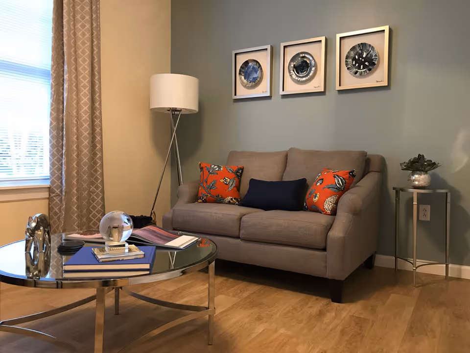 A cozy living room with a beige loveseat adorned with two orange floral pillows and one navy blue pillow. Above the loveseat, three framed abstract art pieces hang on a light blue wall. To the left, a tall floor lamp stands beside a window with beige patterned curtains. In front of the loveseat, a round glass coffee table holds books, a decorative glass orb, and a small sculpture. To the right, a small side table with a potted plant sits against the wall.