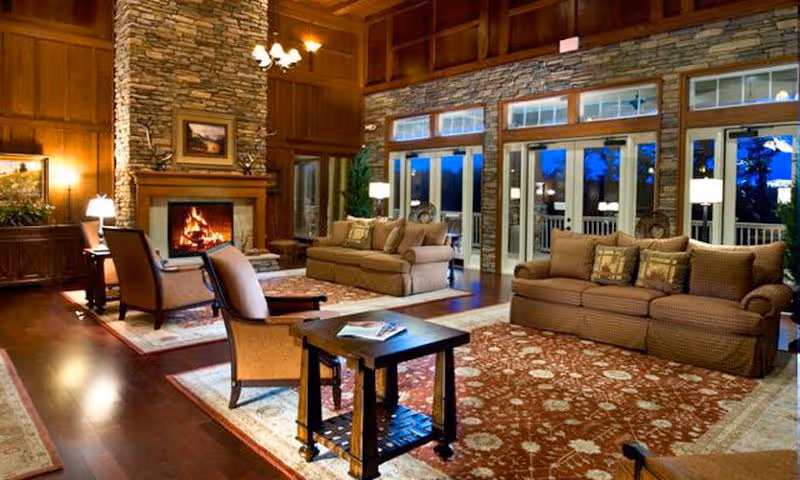 A spacious and warmly lit living room with wooden floors, two large patterned sofas, two armchairs, a wooden coffee table, and a stone fireplace with a fire burning. The room features large windows and glass doors with views of the outdoors, and decorative rugs covering the floor.