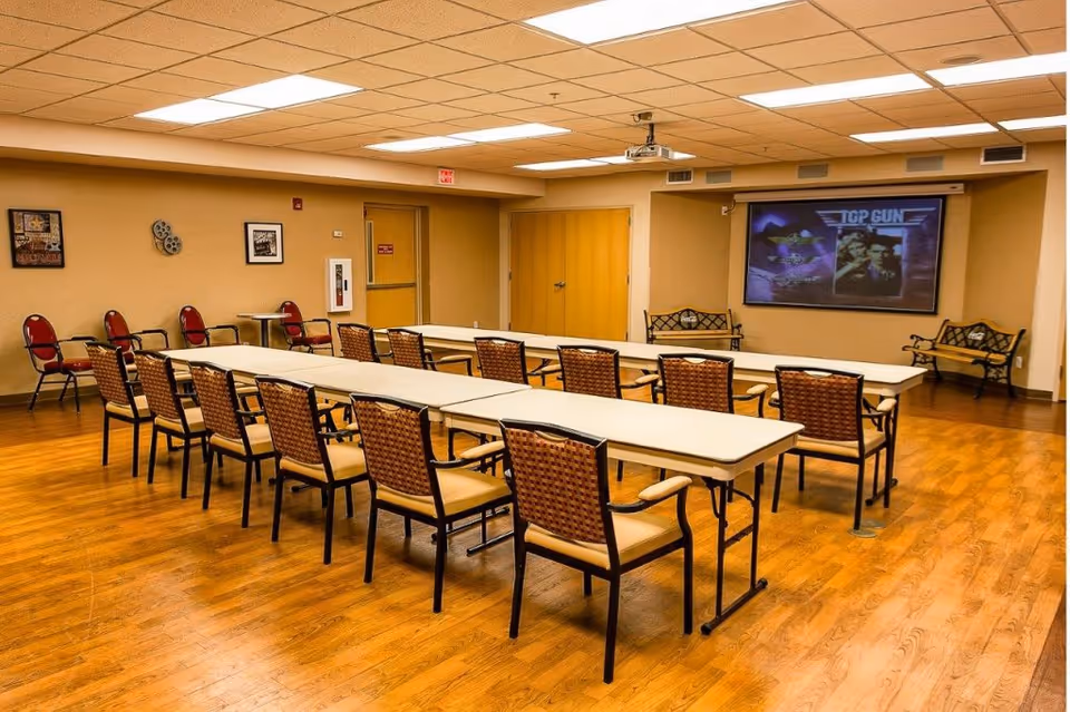 A meeting or activity room with two long tables arranged in a U-shape surrounded by chairs. The room has wood flooring, beige walls, and a ceiling with fluorescent lights. On one wall, there is a large screen displaying the movie Top Gun. There are also some framed pictures and decorative items on the walls, and a couple of benches along the back wall.