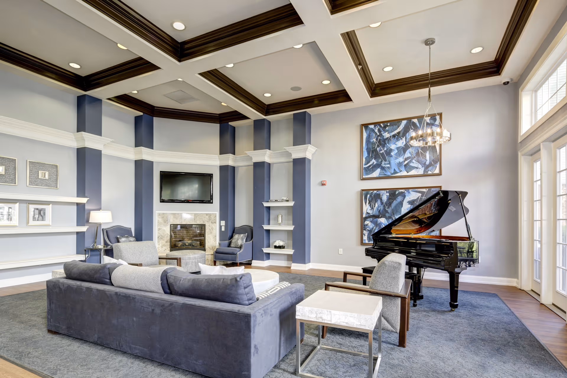 A spacious and elegant living room with a dark blue and gray color scheme, featuring a large dark gray sofa, two gray armchairs, a marble fireplace with a mounted TV above it, a black grand piano, two abstract blue paintings on the wall, and a chandelier hanging from a coffered ceiling. The room has large windows allowing natural light to enter.