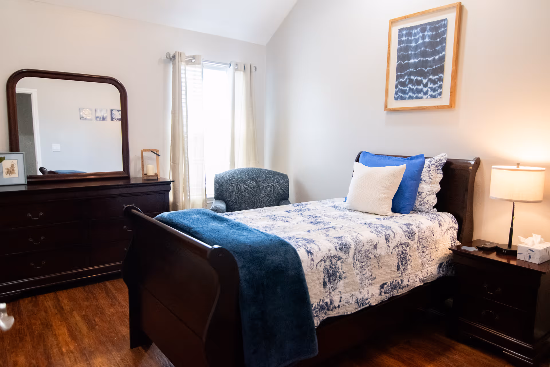 A cozy bedroom with a wooden bed featuring a white and blue patterned quilt and blue and white pillows. There is a wooden nightstand with a lamp and tissue box on the right side of the bed. A wooden dresser with a large mirror is on the left side of the room. A blue upholstered armchair is positioned near a window with light-colored curtains. A framed abstract artwork hangs on the wall above the bed.