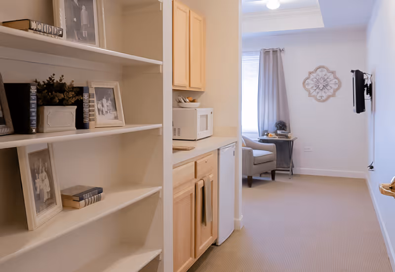 Interior view of a senior living facility room showing a small kitchenette with light wood cabinets, a microwave, and a mini fridge. To the left, there is a white bookshelf with framed black and white photos, books, and a small plant. In the background, there is a sitting area with a gray armchair, a small side table with books and a decorative item, a window with gray curtains, and a wall-mounted TV on the right wall.