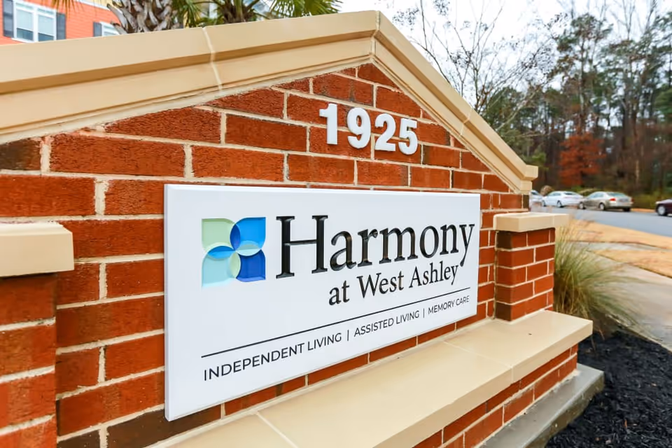 A brick sign for Harmony at West Ashley senior living facility with the address number 1925 displayed above. The sign includes the facility's logo and mentions Independent Living, Assisted Living, and Memory Care services. The background shows a parking lot and some trees.