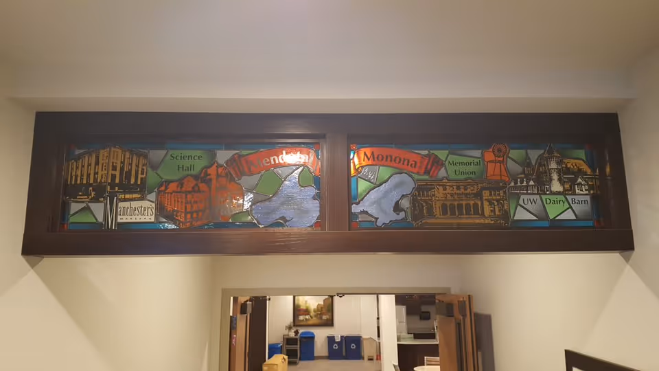 A stained glass window panel above a doorway depicting various landmarks and locations including Science Hall, Memorial Union, UW Dairy Barn, and Manchester's, with colorful geometric background patterns.